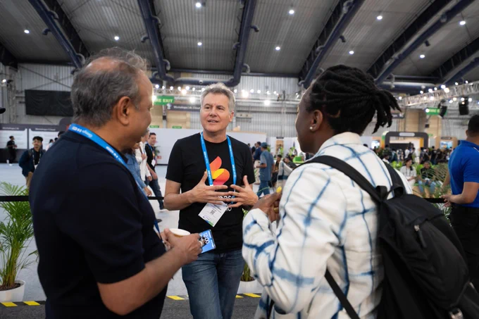 Three attendees are engaged in conversation at a large indoor conference venue. In the center, a smiling person with grey hair wears a black t-shirt with a red and orange Firebase logo, blue jeans, and a Google lanyard with an ID badge identifying "Angel Calvo", gesturing while speaking. To the left, a person with dark hair wears a dark blue shirt and a Google lanyard; to the right, a person with dark braided hair and yellow-painted fingernails wears a white and blue patterned jacket and a black backpack, listening attentively. The background shows blurred conference attendees, various booths, a "FIRE EXIT" sign, and "Workshops | AI, Cloud" faintly visible on a distant sign.