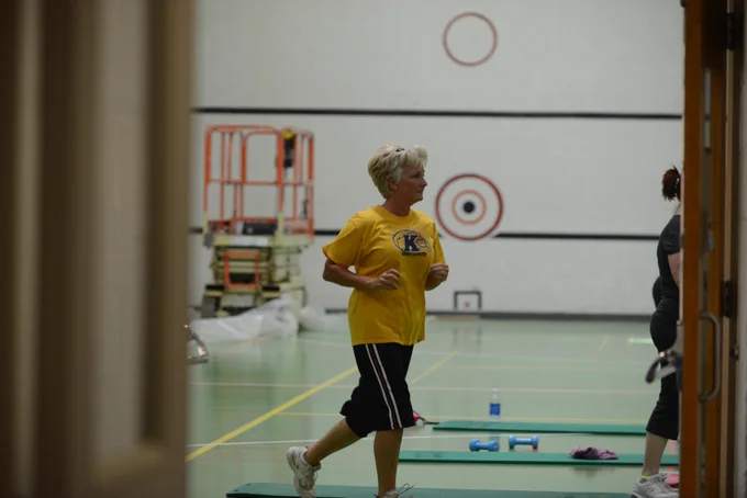 Woman in a yellow Kent State t-shirt walks in a gym during a fitness class, with exercise mats and weights on the floor.