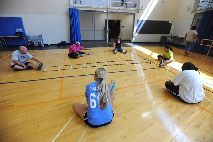 Group of adults sitting on a gym floor stretching near an agility ladder during a fitness class.