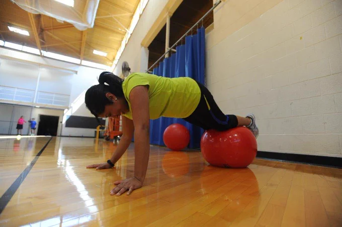 Woman in a green shirt performs a plank with her feet on a red exercise ball in a brightly lit gym.