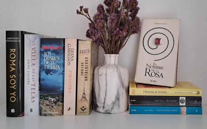 Fotografía de varias novelas históricas: "Roma soy yo" de Santiago Posteguillo, "La villa de las telas" de Anne Jacobs, "Los pilares de la Tierra" de Ken Follett, "Forastera" de Diana Gabaldon, "París" de Edward Rutherford, "El nombre de la rosa" de Umberto Eco", "Memorias de Adriano" de Marguerite Yourcenar, "Yo, Claudio" de Robert Graves y "Capitán de mar y guerra" de Patrick O'Brian. Junto a los libros hay un jarrón con flores secas.