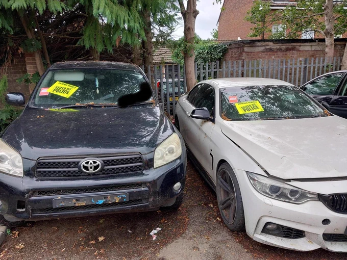 This image shows a white BMW and a Black Toyota with a yellow sticker, stating 'Untaxed vehicle seized by police'