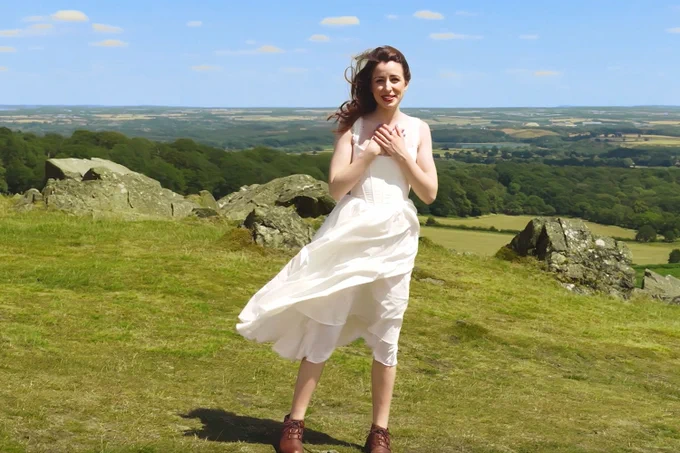 A photo showing a woman in a white dress singing to camera, with rolling hills behind her 