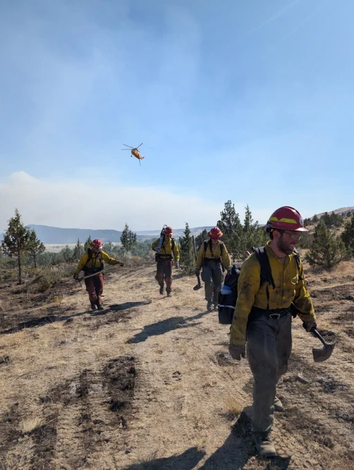 Firefighters hiking down a trail with a helicopter in the background.