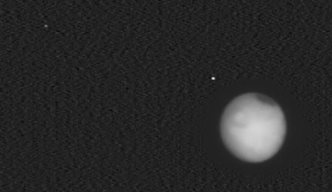 Black-and-white image of the disc of Mars against the darkness of space. Two small dots to the left of the planet are two of its moons.