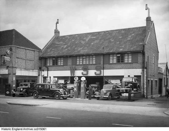Black and white photograph looking across an road towards a service station with petrol pumps and cars in the forecourt. The domestic-looking building has two storeys, a pitched roof and gable ends. A young man in a boiler suit stands next to a petrol pump.