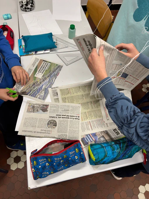 Des élèves travaillent en groupe autour d’une table recouverte de feuilles de papier journal. On aperçoit des ciseaux, des règles et des trousses. Ils manipulent le papier pour tester des structures, probablement dans le cadre d’une maquette ou d’une étude sur les matériaux.