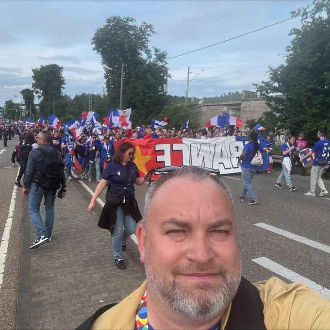 A selfie of a man in front of a lively street march of French football fans carrying flags and a large “FRANCE” banner, creating a festive and patriotic atmosphere during UEFA Women’s EURO 2025.
