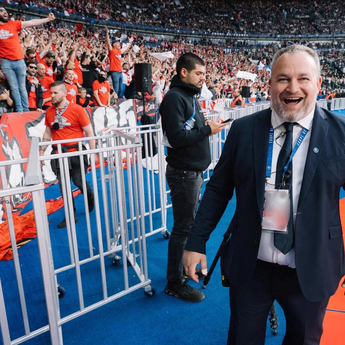 A smiling SLO stands pitchside in front of a passionate crowd of fans wearing red shirts, with security and stewards in the background during a high-energy international football match.
