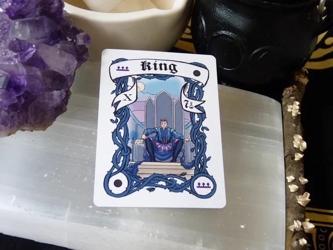 The New Moon King card has a new moon in 2 corners and a purple crown in the other two, with blue border vines framing a Caucasian man with a crown on a throne, in blue robes, stamping a scroll with his ring. In the banner there is the Roman Numeral X on the left side and the Arabic number 7 on the right.  The card is laying on a selenite slab, surrounded by a cluster of purple amethyst, a white mortar and pestle, a triple moon goddess cauldron, and a stick with lichen, all on a black and yellow cloth.