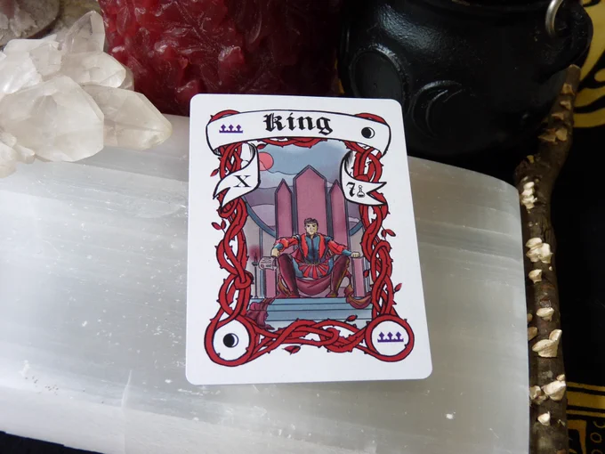 The Crescent Moon King card has a crescent moon in 2 corners and a purple crown in the other two, with red border vines framing a Hispanic man with a crown on a throne, in red robes, stamping a scroll with his ring. In the banner there is the Roman Numeral X on the left side and the Arabic number 7 on the right. The card is laying on a selenite slab, surrounded by a cluster of clear quartz, a red candle, a triple moon goddess cauldron, and a stick with lichen, all on a black and yellow cloth.