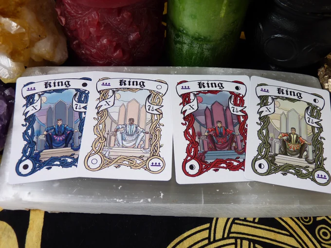 All 4 King cards are laying on a selenite slab in order of New, Full, Crescent, and Quarter, surrounded by a cluster of purple amethyst, a cluster of yellow citrine, a red candle, a green candle, a triple moon goddess cauldron, and a stick with lichen, all on a black and yellow cloth.