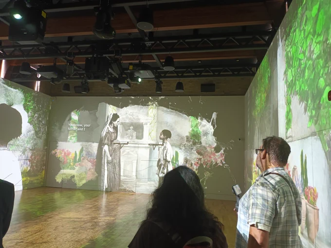 A room with two people with their backs to image, an image of a woman and a boy are on one of the walls.
