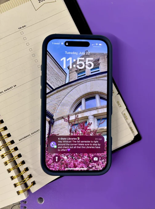 Smartphone displaying a lock screen with a large digital clock, a calendar notification for "Tuesday, July 22," and a text message from "K-State Libraries" about the upcoming semester. The background image on the phone shows a brick building with arches and cherry blossoms. The phone rests on an open planner with some handwritten notes.