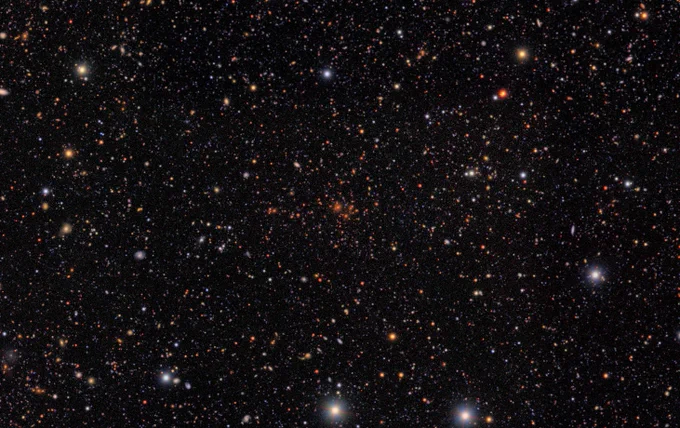 A glittering tapestry of thousands of galaxies in varying sizes, shapes, and colors. At center is a distinct collection of galaxies, all of a similar red color.