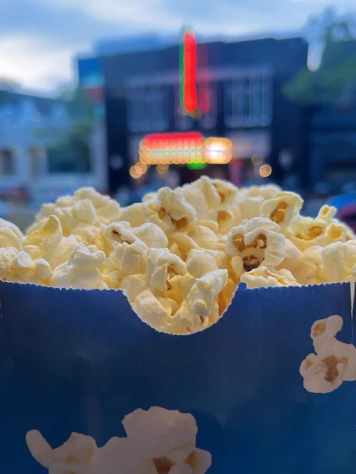 Closeup of popcorn in a blue bag in focus with the neon lights of a movie theater, The Little, in the background 
