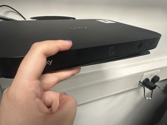 Close-up of a hand holding a black Sky box. A white shelf and electrical outlet are visible in the background.