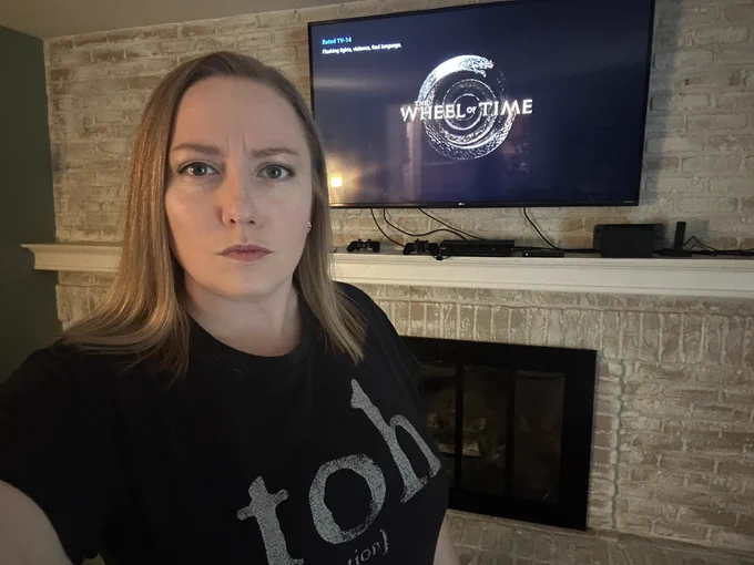 Woman with a grumpy face is wearing a black t-shirt with the words toh (obligation) on the front. Behind her is a tv showing The Wheel of Time show logo; a bronze snake coiled upon itself with the words “The Wheel of Time” across it.