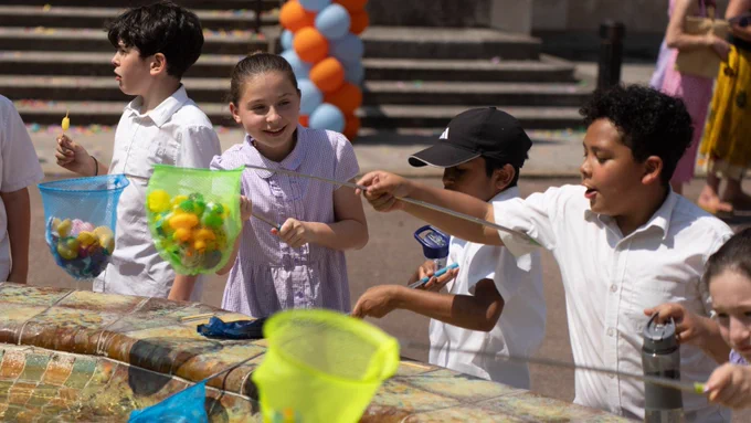A group of children taking part in an outdoor activity using fishing nets. They are gathered around a circular stone fountain, catching items floating in the water with colourful nets. The children are dressed in light summer clothes, and the background features steps decorated with orange and blue balloons.