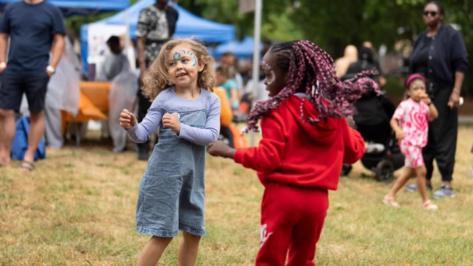 Two children dancing or playing on a grassy area during an outdoor event. One child wears a blue dress over a long-sleeved shirt, and the other is dressed in a red outfit. In the background, people are standing or sitting near blue tents and tables.