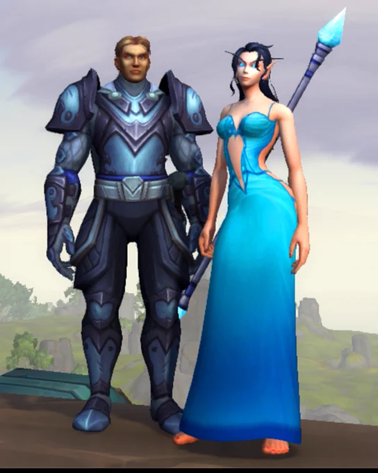 In World of Warcraft, Michael Allen’s human paladin stands beside Cindi Curtis’s Void Elf mage on the wall of a city with soft clouds and rolling hills in the background. He wears full dark-steel armor with glowing blue highlights, while she’s dressed in an elegant sky-blue transmog gown that shows off her silhouette. Her glowing blue staff rests against her shoulder.