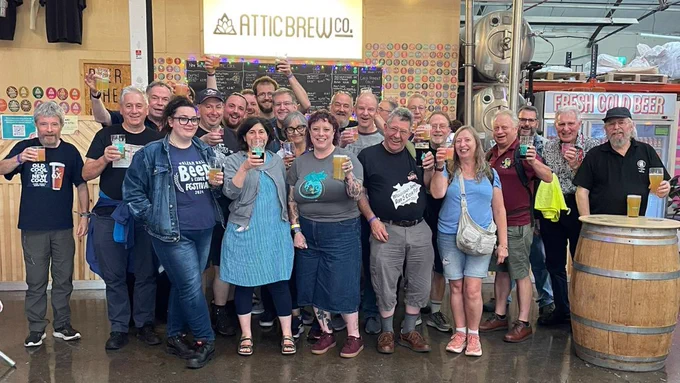 A line of people cheering next to a barrel and a wooden background with the Attic Brew co logo behind