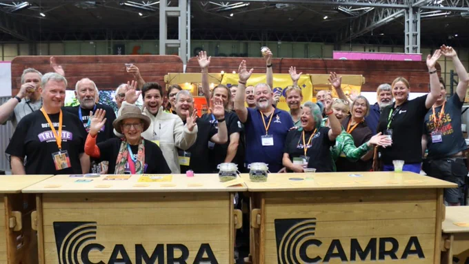 A long line of people cheering behind wooden bars with the black CAMRA logo on them.
