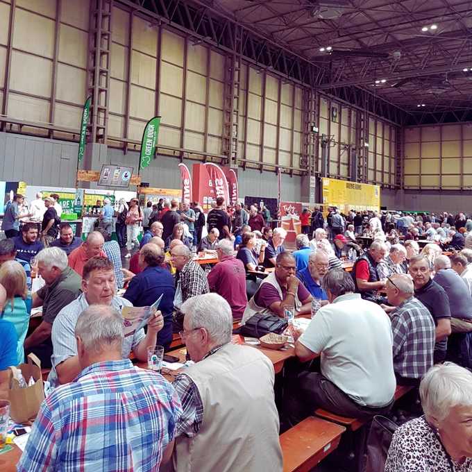 A large crown of people sat at wooden benches inside a large hall. There are banners  in the distance.