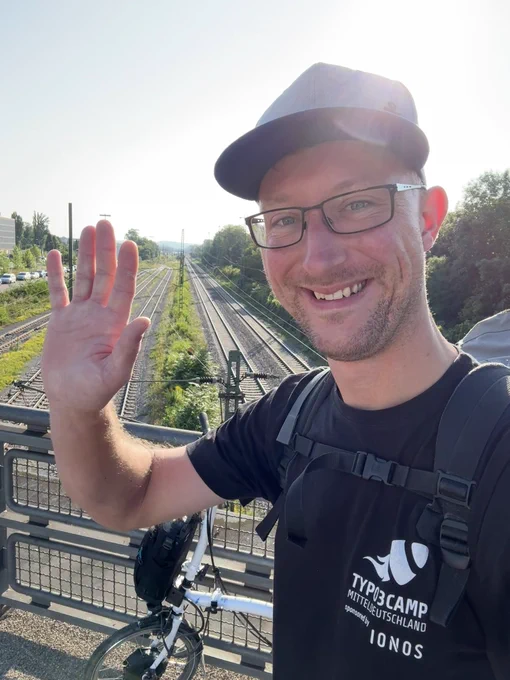 I'm standing on a bridge in the sun, waving goodbye from Karlsruhe after the TYPO3 Developer Days 2025, with the railway tracks and my folding bike behind me.