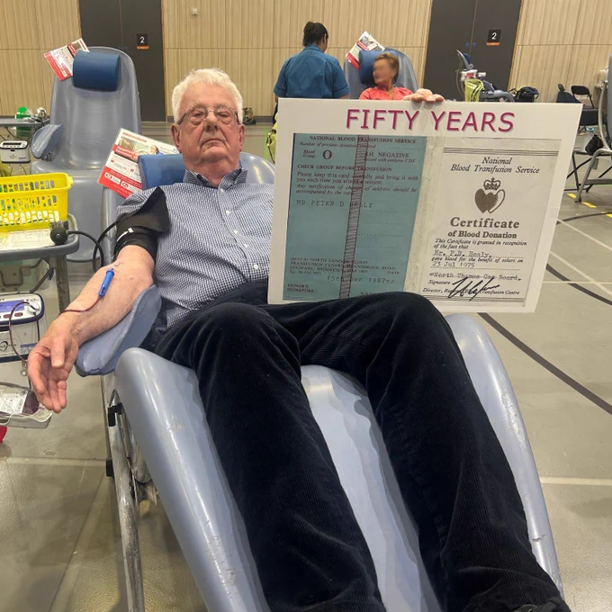Pete sits in a donor chair while giving blood. He holds up a large version of his donor card from 1975.