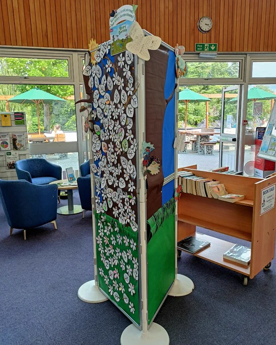 Photograph of paper mural depicting a large brown tree, green grass at the bottom, and a small sun in the top left corner on either side of a tall triangular frame. The frame is decorated with paper butterflies of various sizes. One side of the mural is covered in paper flowers and leaves, with each one representing a different child participation in the Summer Reading Challenge. The other side is bare waiting to be populated with leaves and flowers. 