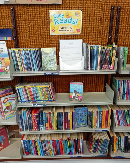 Photograph of a row of shelves under a sign that reads "Easy Reads!". The books on the top two shelves have yellow dot stickers on the spine denoting them as part of the Easy Reads section. In the middle of the top shelf are a set of A4 leaflets with information on dyslexia and dyslexia friendly books.