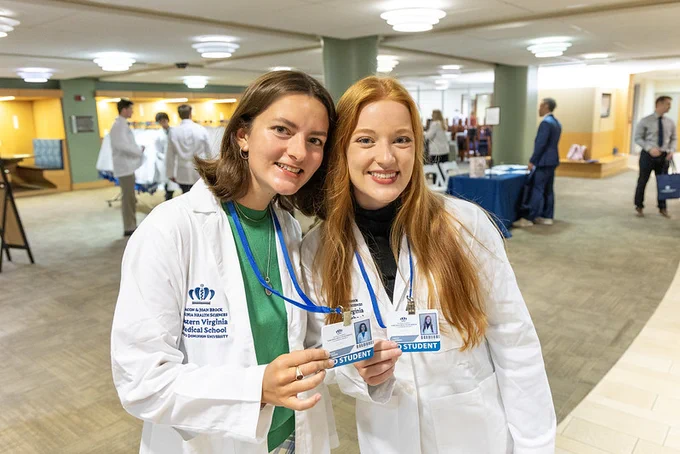 Two individuals in white lab coats smiling and holding identification cards. 