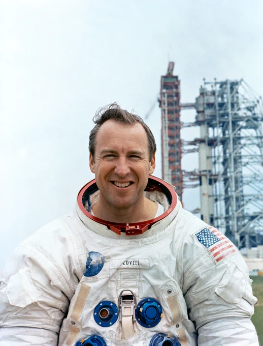 Astronaut Jim Lovell, wearing a white spacesuit with no helmet, stands outside with a tall rocket and launch tower behind his left shoulder. Lovell is pictured from the chest up; he is looking at the camera and smiling. Credit: NASA