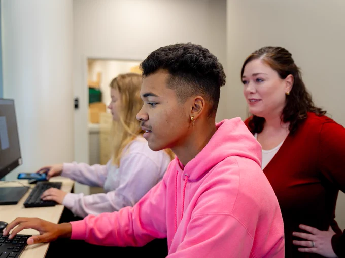 A person in a pink sweatshirt working on a computer in a classroom setting, with two other individuals focused on their screens in the background. Another individual is standing behind, observing the screen.