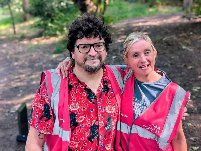 Kelly and a man stand together in the park, both wearing parkrun volunteer hi-vis vests. Kelly has her arm around him and is smiling, while he’s wearing glasses and a bright red floral shirt. The background is leafy and green.