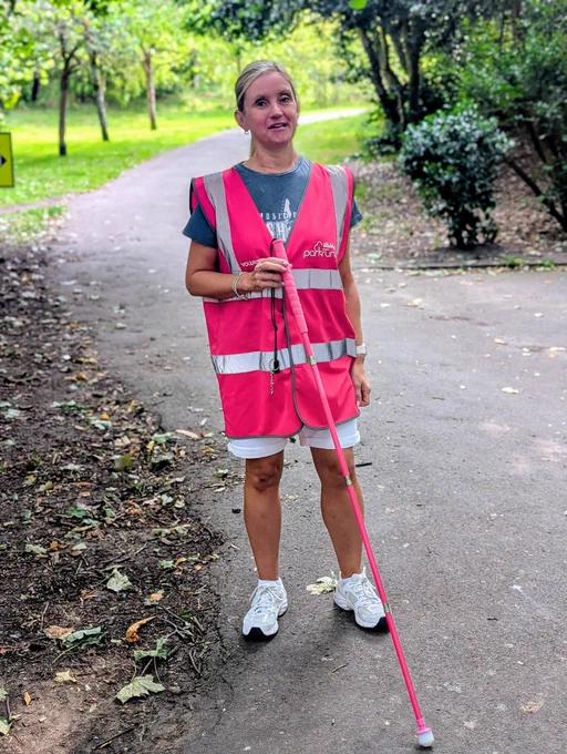 Kelly standing on a park path surrounded by trees, wearing a bright pink parkrun volunteer hi-vis vest over a grey T-shirt and white shorts. She’s holding a matching pink long cane, smiling towards the camera