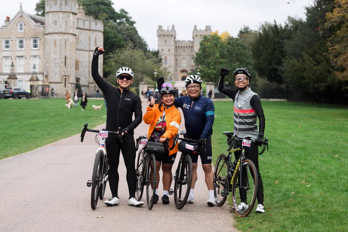 Four smiling cyclists pose with their bikes near Windsor Castle, raising their arms in celebration. The castle and trees provide a scenic backdrop.