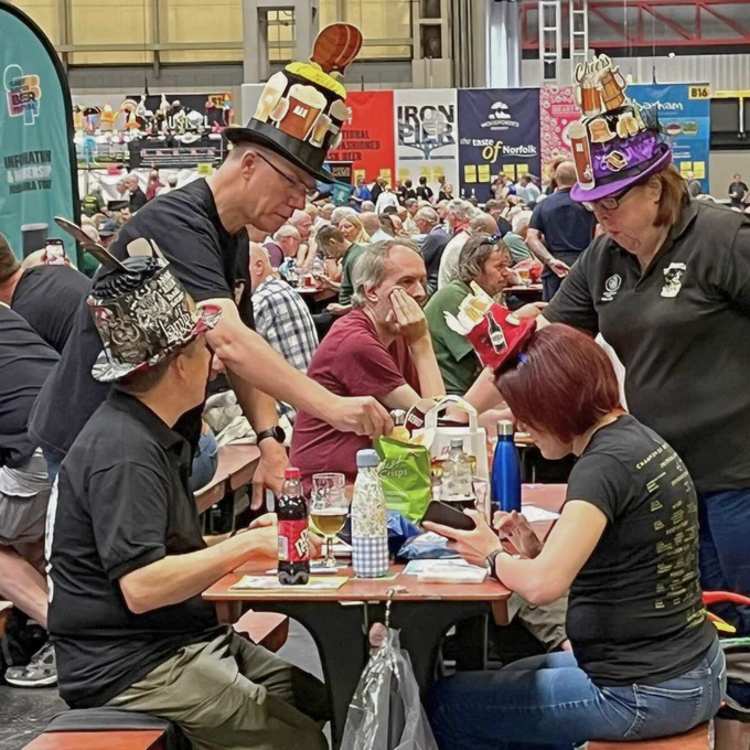 A group of people wearing novelty beer hats sat on a wooden bench with bottles, bags and glasses on top.