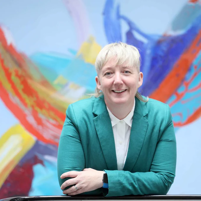 
A blonde woman with a short haircut smiles at the camera. She is wearing a dark teal blazer over a white shirt and is leaning forward with her arms crossed on a dark surface. Behind her is a light blue wall with a large, colorful abstract painting in shades of red, orange, and blue.