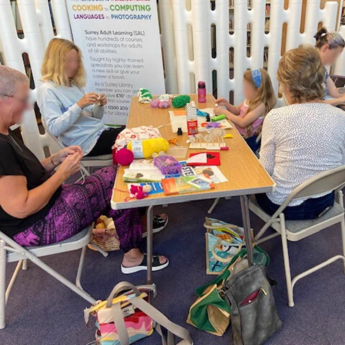 Photograph of a group of adults and one child sitting at a long wooden table doing a variety of different crafts. On the table, there are coloured balls of wool and other various art supplies.