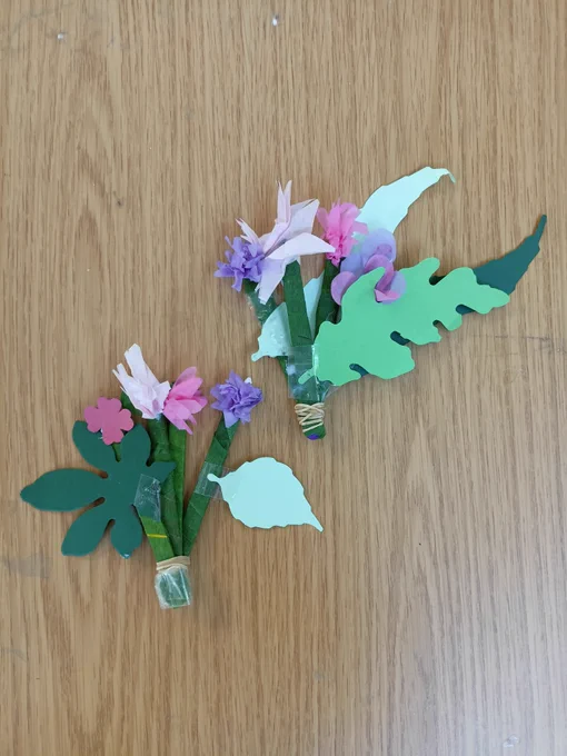 Photograph of two bouquets of crafted flowers on a wooden table. The stems are green lollipop sticks that are tied together at the bottom, with large paper leaves in different shades of green and crushed tissue paper flowers in shades of pink and purple.