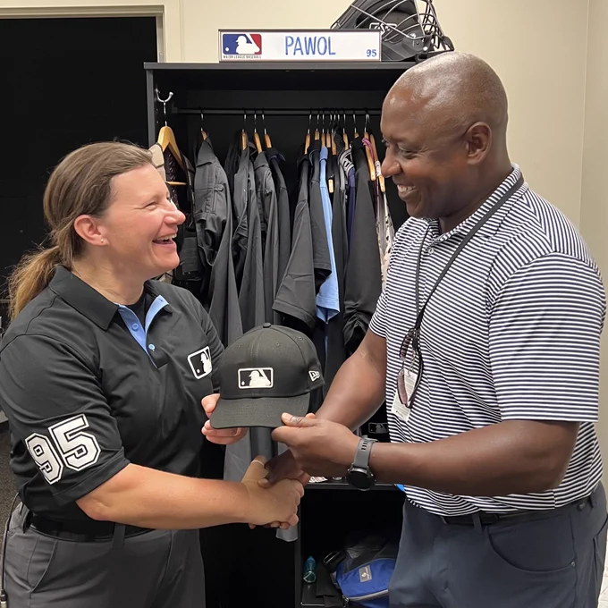 Jen Pawol, left, stands at her locker and shakes hands with Michael Hill, Major League Baseball Senior Vice President of On-Field Operations and Workforce Development. Both are holding the black MLB cap worn by Pawol on August 9, 2025, when she became the first woman to umpire in a regular-season major league game.