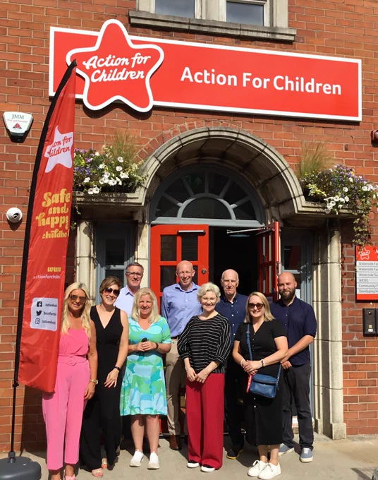 Image shows Bronagh (front right in black dress) pictured during a recent NI Committee visit to the Action for Children offices in Ebrington. It’s a really sunny day and the group of nine people are standing at the front door which has a large red Action for Children sign above it.