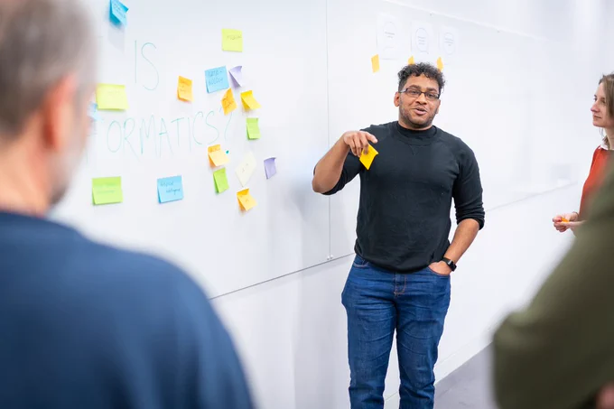 A man in a black jumper stands in front of a white board covered in brightly coloured Post-it notes. The notes surround the text 'What is bioinformatics?' 
