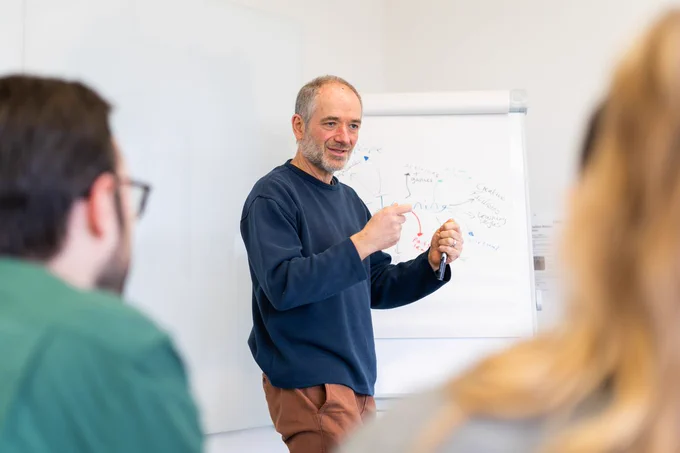 A man in a blue jumper stands in front of an easel and a flipchart holding a marker pen. 