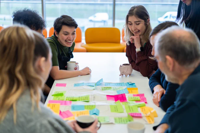 A group of smiling people sit at a large white table covered in bright coloured post-it notes. Some of the people are holding coffee cups.