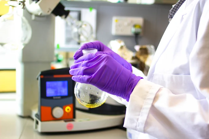 Scientist wearing purple gloves holds a glass flask in a laboratory, with scientific equipment visible in the background.