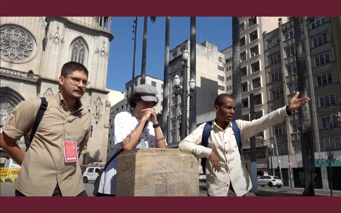 Three people, one gesturing, stand in front of the São Paulo Cathedral in a busy urban setting taken from the short film 'Rhythms of Negotiation'.