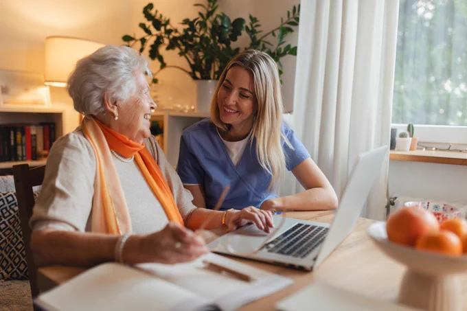 Elderly woman and healthcare worker sitting at a table using a laptop in a cozy, well-lit room with plants and books.
